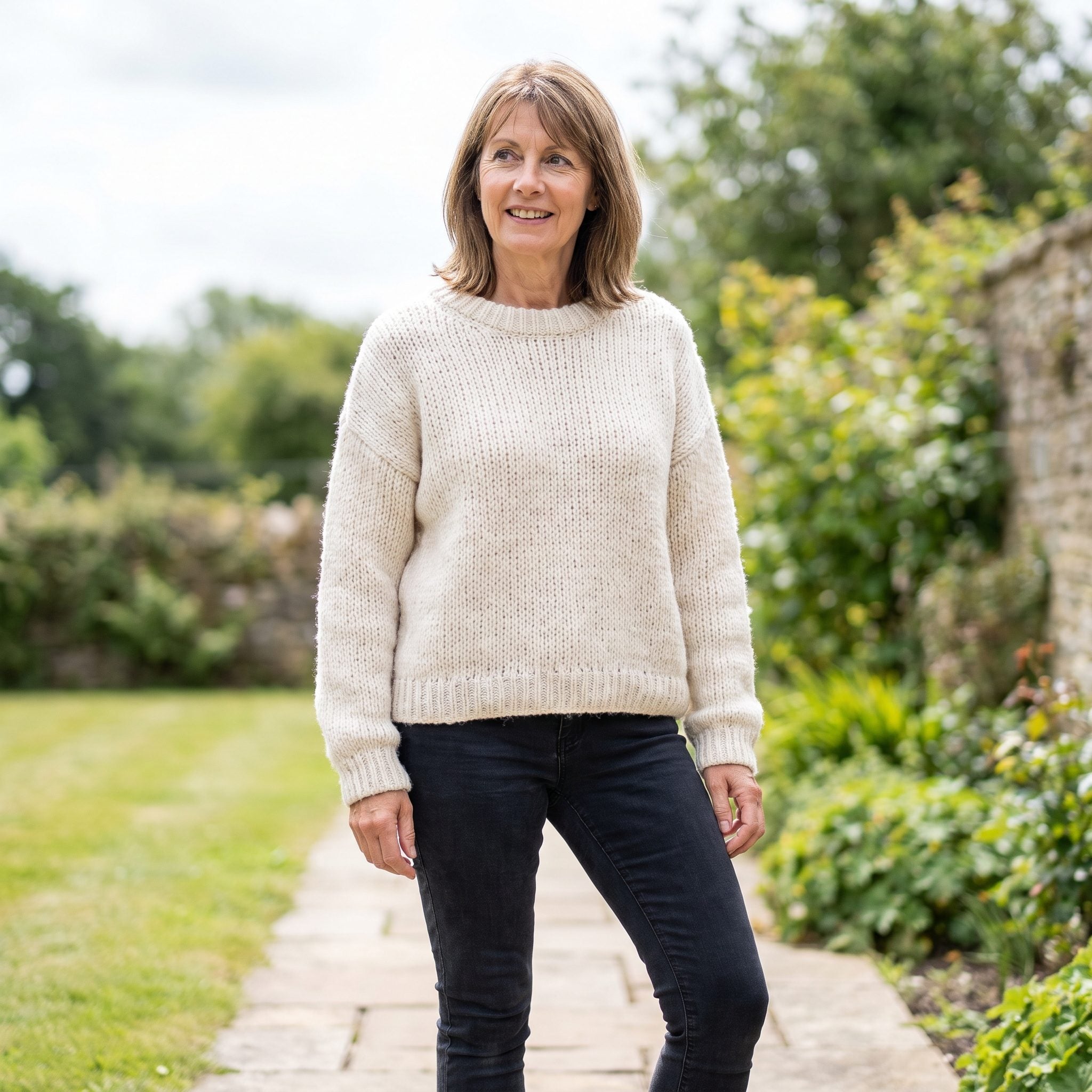 Woman in deep-blue skinny jeans and a thick cream cable-knit sweater with a straight hem cutting across the hip