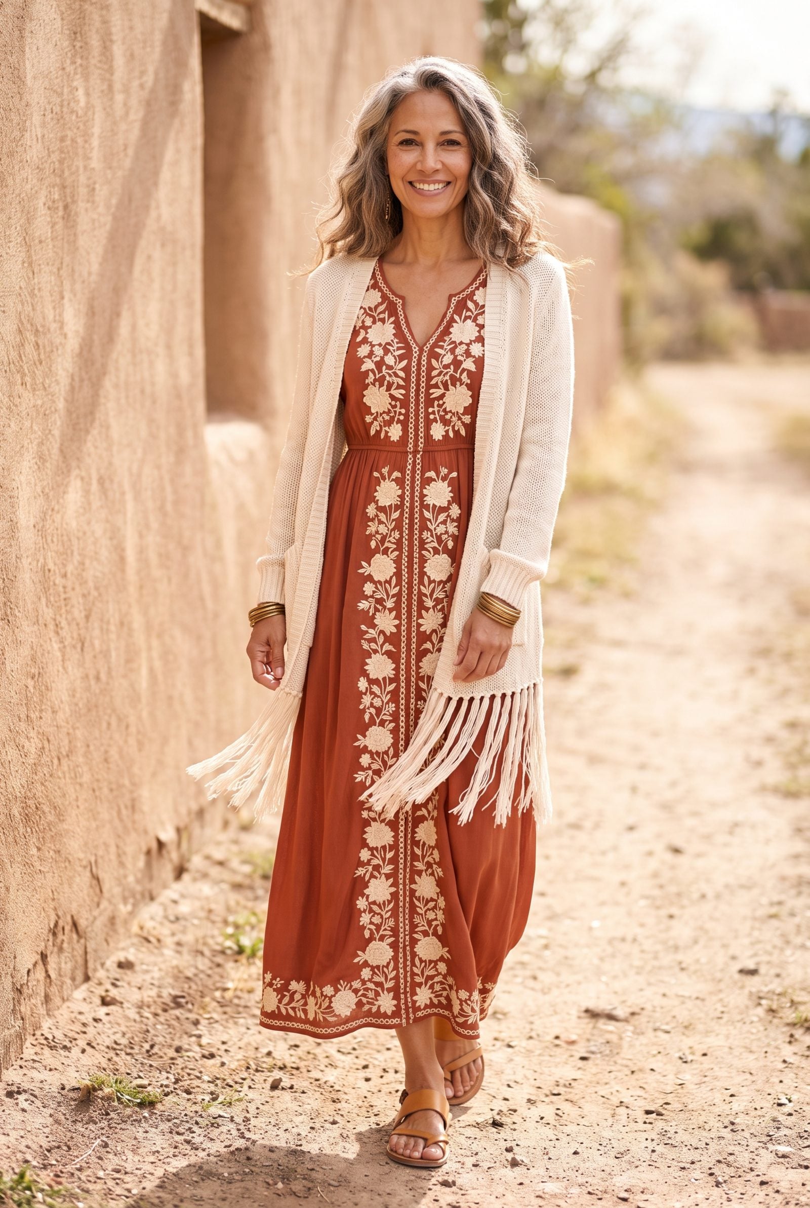 Woman in an embroidered rust boho maxi dress under a cream fringed cardigan, fringe overlapping the embroidered panel.