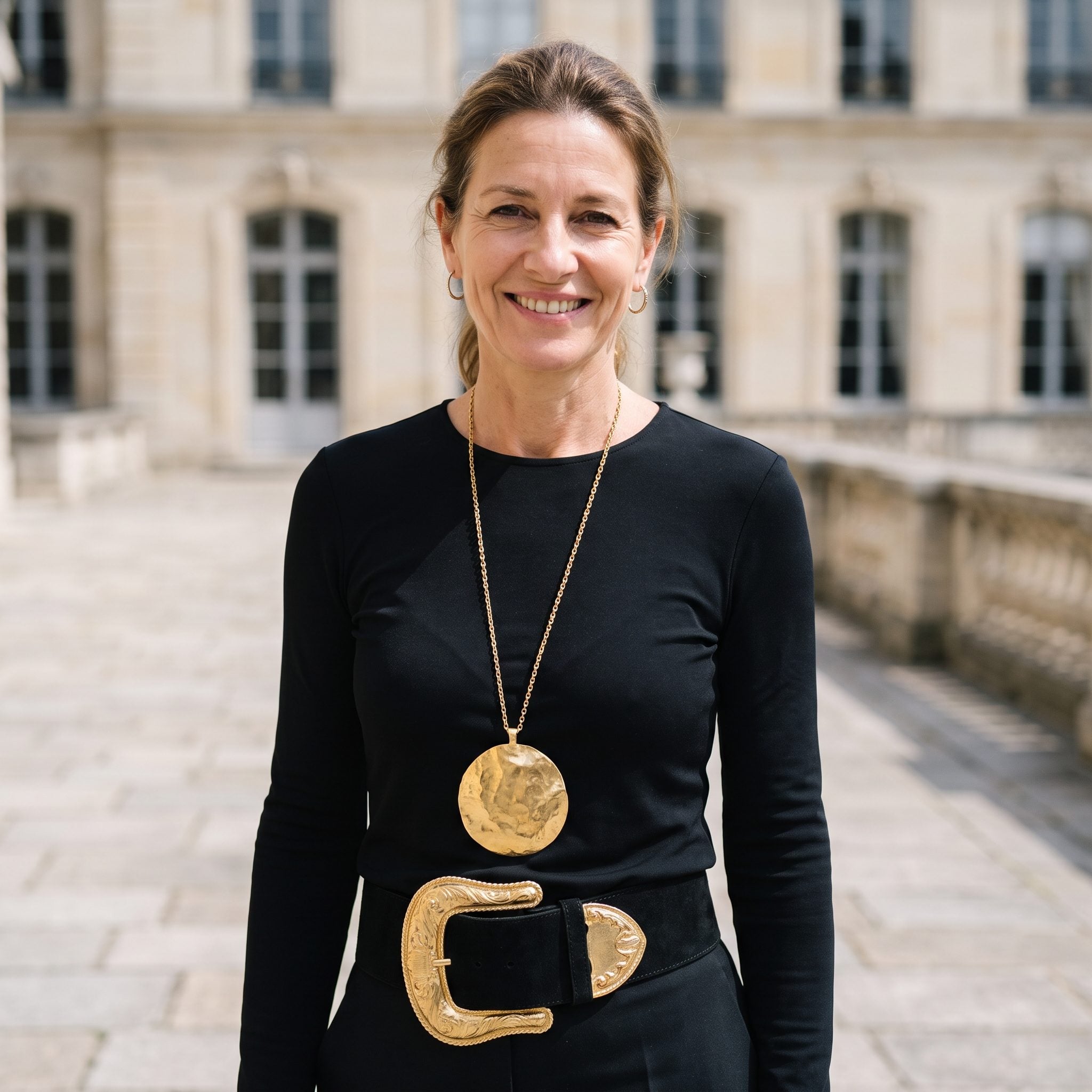Woman in a black blouse with a long pendant necklace ending just above a large ornate gold belt buckle.