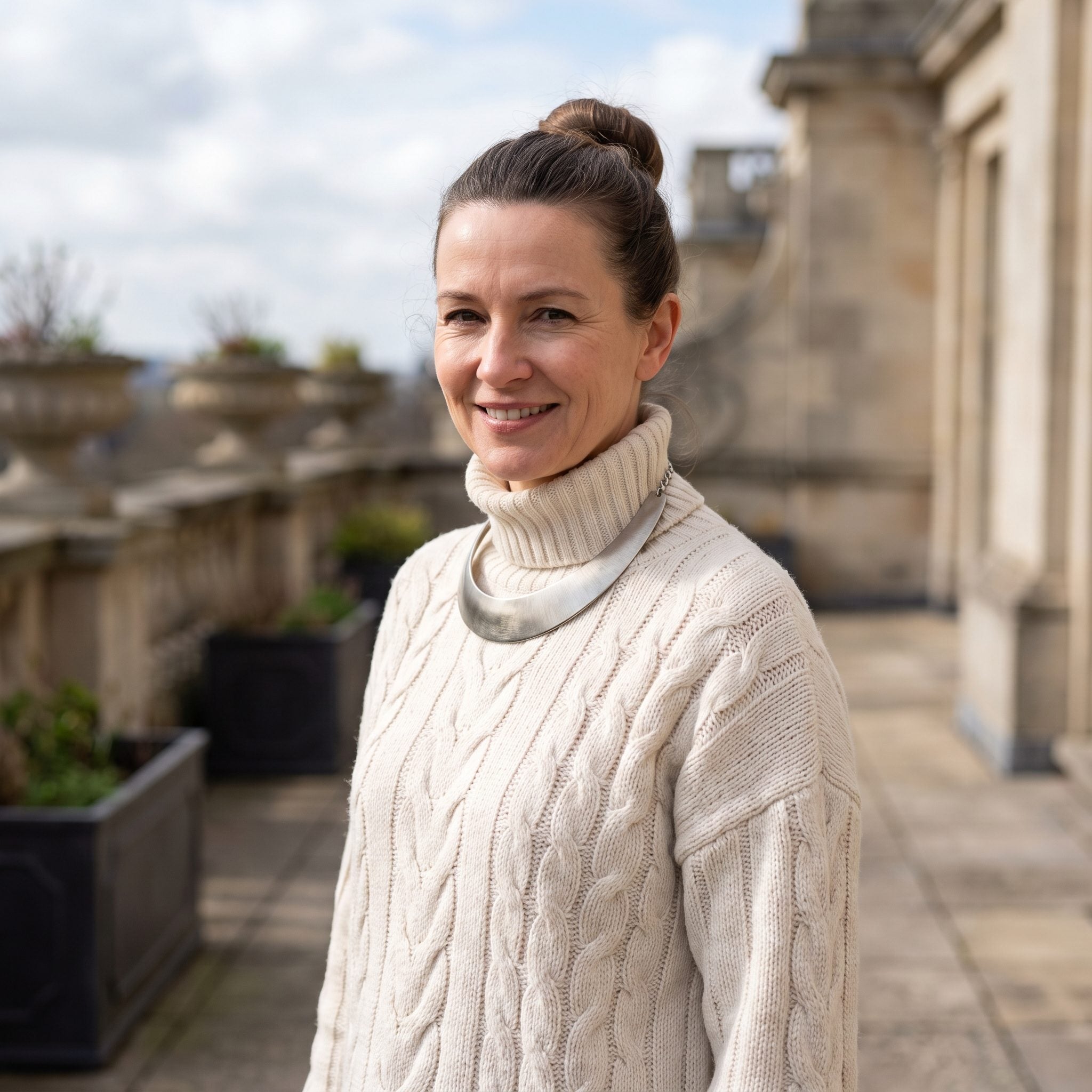 Woman in a cream cable-knit turtleneck with a rigid collar necklace crowded beneath the thick knit collar.