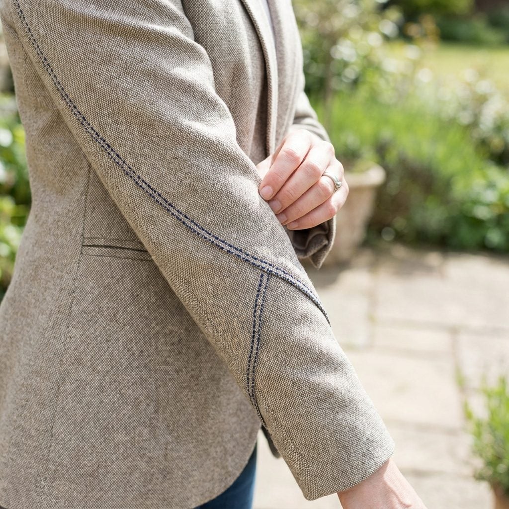 Woman in a camel blazer and indigo jeans, blazer sleeve seams twisting around her arms with spiral folds.