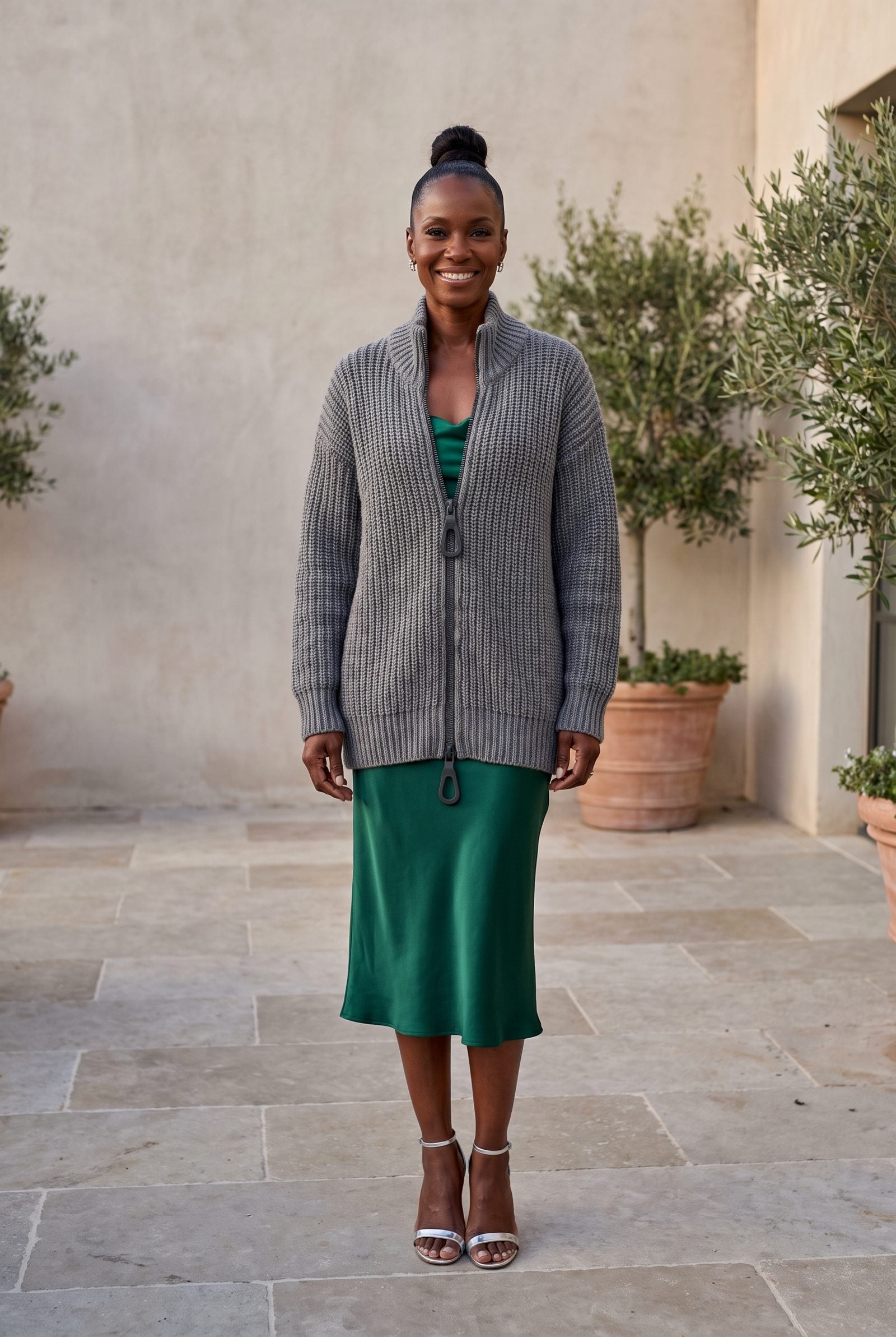 Woman in an emerald satin cocktail dress under a fully zipped grey chunky knit cardigan flattening the satin drape.