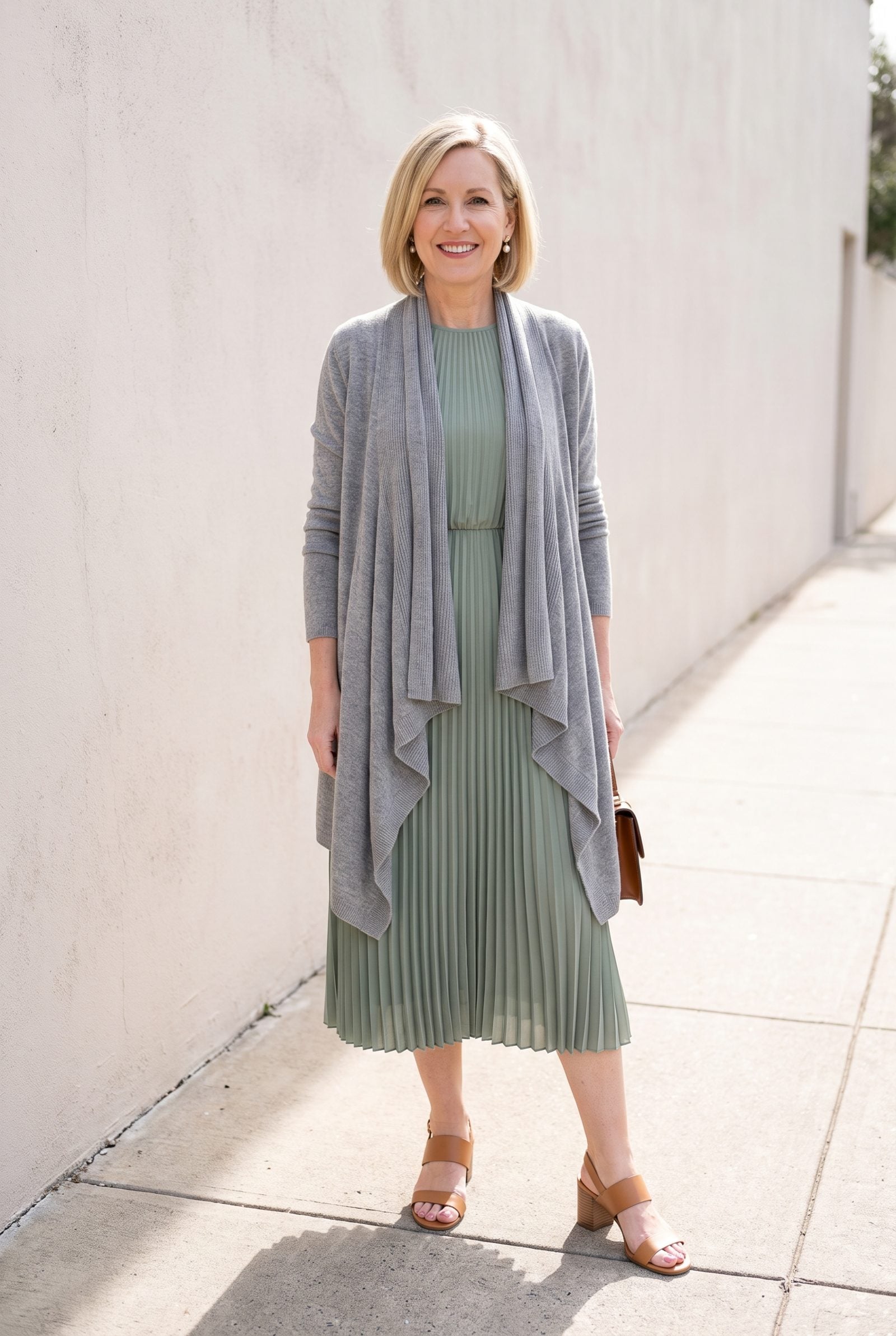 Woman in a sage pleated midi dress under an open grey waterfall cardigan whose draped front folds cover the pleats down the
