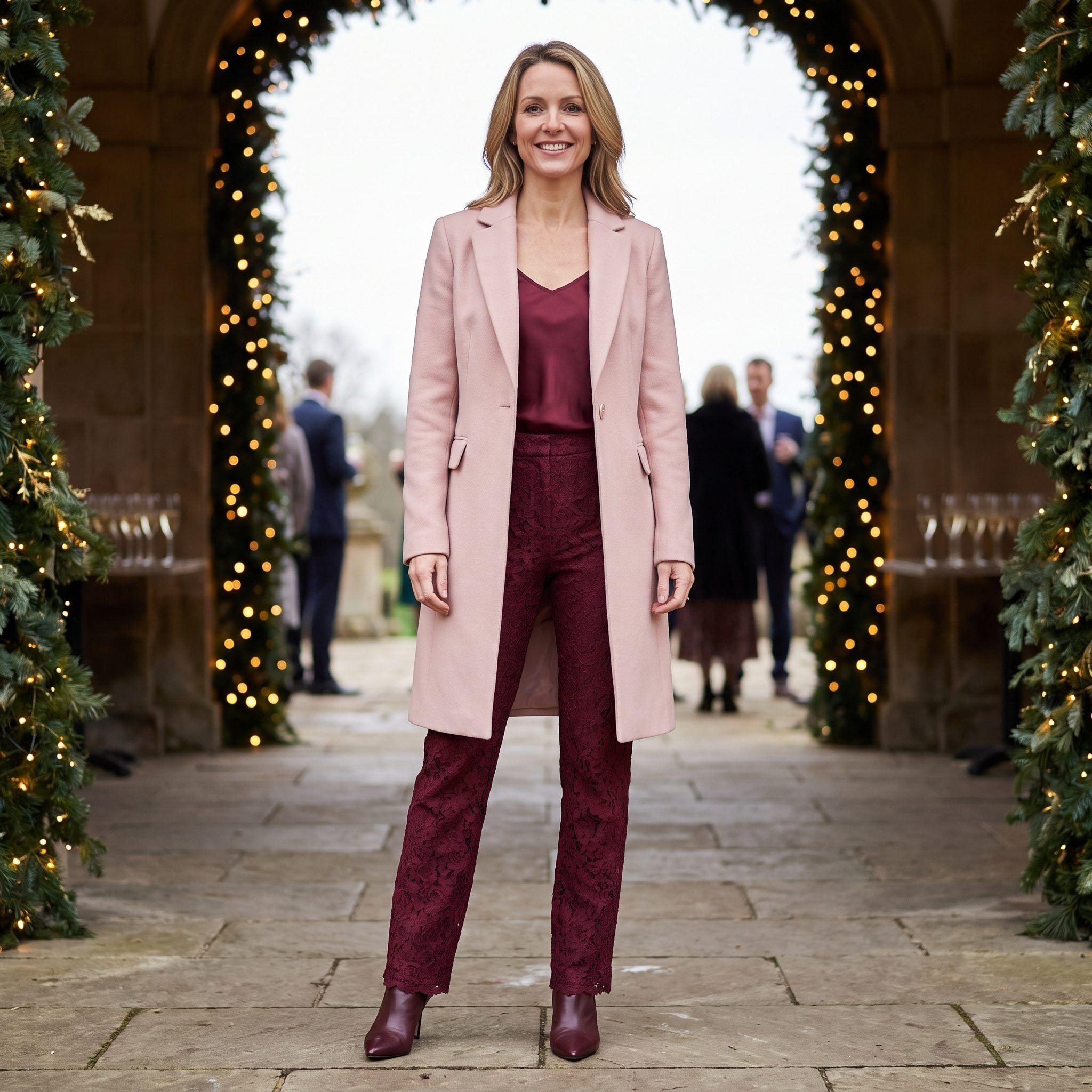 Woman in burgundy lace trousers and camisole under an open blush mid-calf wool coat with burgundy ankle boots